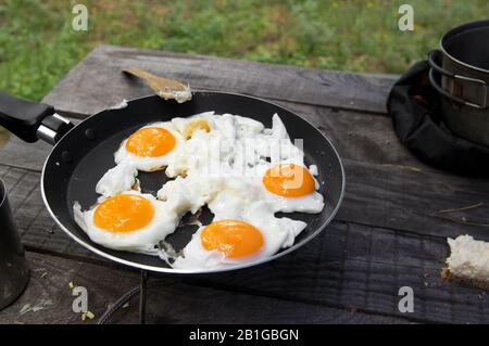 Uova fritte cucinate in padella sulla stufa a gas da campeggio su una vecchia tavola di legno. Cucina all'aperto nella foresta. Foto Stock