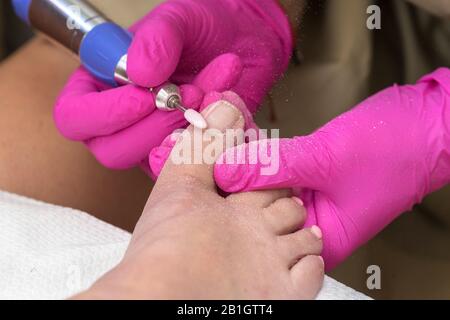 Il Processo Di Pedicure. Pedicurist donna rimuove il vecchio gel polacco dalle unghie delle dita dei piedi utilizzando un trapano elettrico per unghie. Mani femminili con speciale gomma rosa Foto Stock