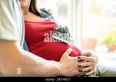 Mamma giovane donna che tiene mano padre sul suo addome con le scarpe bambino sul ventre - Famiglia in attesa di bambino con luminoso sullo sfondo - Incinta e. Foto Stock