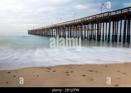 Molo di Ventura, California; vista dalla spiaggia, che si estende in acqua. Oceano verde, cielo nuvoloso. Foto Stock