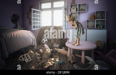 Ragazza bionda caucasica di 3 anni spaventata e in piedi su un piccolo tavolo vede il fango entrare nella sua camera da letto. Concetto di disastro e problema di famiglia. Foto Stock