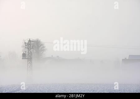 Elettricità pilone ai margini del villaggio in inverno, nevoso nebbioso paesaggio rurale alla luce del mattino, concetto per sognante, mistico e agricolo Foto Stock