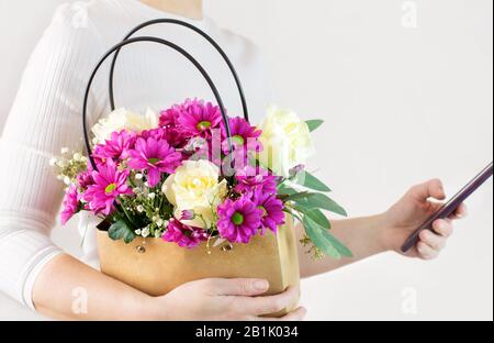 Una donna tiene un cesto con fiori e uno smartphone nelle sue mani. Floriestry, consegna da un negozio di fiori. Primo piano. Spazio di copia. Foto Stock