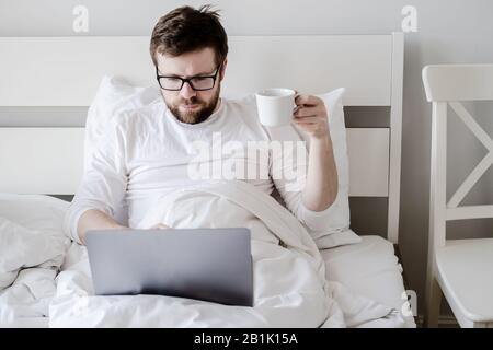 L'uomo focalizzato, con una tazza di caffè in mano, lavora su un computer portatile, seduto a letto, sotto luce naturale. Moderna vita. Foto Stock