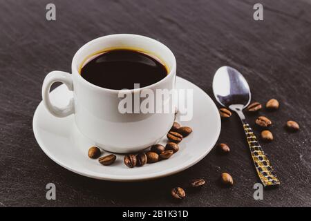 Una tazza bianca e un piattino contenente caffè nero senza latte, un cucchiaio accanto ad esso e chicchi di caffè marrone sono sparsi. Foto Stock