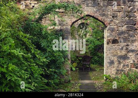 Rustica antica arcata in pietra in un giardino appartato e sovracresciuto con piante da arrampicata e tavoli e sedie Foto Stock