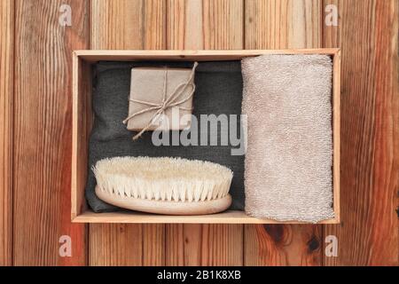 Sapone fatto a mano avvolto in carta artigianale, asciugamano e spazzola massaggio si trovano in una scatola di legno su uno sfondo di legno. Foto Stock