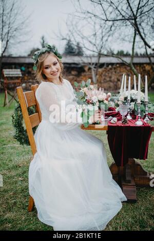 La sposa si siede vicino al tavolo da banchetto con candele in candelieri, piatti, bicchieri per vino, una disposizione floreale e una tovaglia rossa. Foto Stock