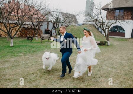 I newlyweds hanno un divertimento e corrono sul prato accanto al cane Samoyed sullo sfondo della casa. Foto Stock