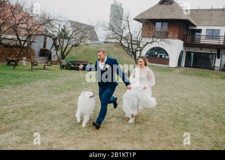 I newlyweds hanno un divertimento e corrono sul prato accanto al cane Samoyed sullo sfondo della casa. Foto Stock