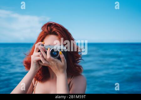 Persona all'oceano con macchina fotografica. Bella donna capelli rossi in spiaggia con fotocamera. Lezioni di fotografia Foto Stock