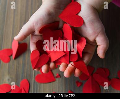 Cuori rossi di carta in bambini mani sullo sfondo di legno. Un regalo per il giorno di San Valentino e il giorno di madri. Foto Stock