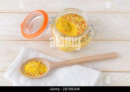 Condimento universale in un vaso di vetro con clip top e in un ampio cucchiaio di legno su un tavolo da cucina in legno bianco. Condimento con sale e verdure secche. Foto Stock