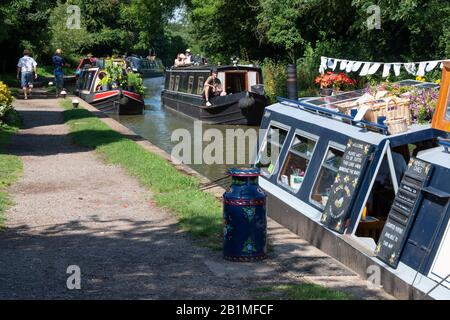 Barca del canale che viaggia lungo il canale di Oxford e che passa un caffè su una chiatta ormeggiata accanto al canale di Braunston, Northamptonshire, Inghilterra Foto Stock