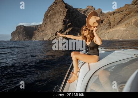 Donna in costume da bagno e cappello da sole che gode di viaggio in oceano, navigando su uno yacht vicino alla costa rocciosa mozzafiato su un tramonto. Concetto di un lusso estate di caccia e di viaggio ricreativo Foto Stock