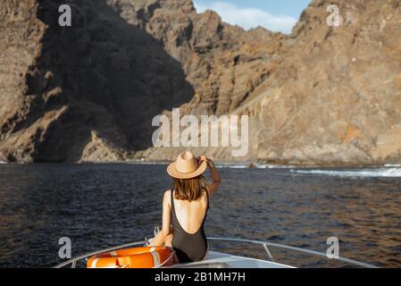 Donna che gode di un viaggio in oceano seduto con la lifebuoy sul naso dello yacht mentre naviga vicino alla costa rocciosa mozzafiato su un tramonto Foto Stock