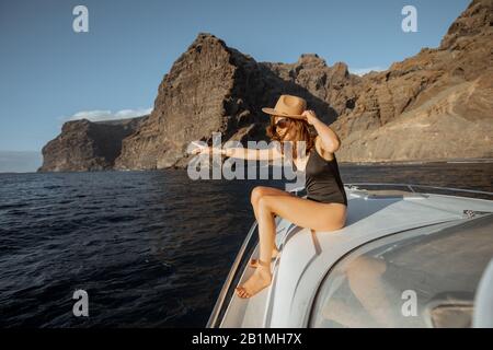 Donna in costume da bagno e cappello da sole che gode di viaggio in oceano, navigando su uno yacht vicino alla costa rocciosa mozzafiato su un tramonto. Concetto di un lusso estate di caccia e di viaggio ricreativo Foto Stock