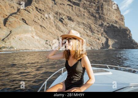 Donna in costume da bagno e cappello da sole che gode di viaggio in oceano, navigando su uno yacht vicino alla costa rocciosa mozzafiato su un tramonto. Concetto di un lusso estate di caccia e di viaggio ricreativo Foto Stock