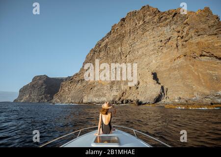 Donna in costume da bagno e cappello da sole che gode di un viaggio sull'oceano seduto sul naso dello yacht mentre naviga vicino alla costa rocciosa mozzafiato su un tramonto. Ampia vista mare Foto Stock