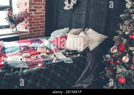 Tazze di caffè e scatola regalo posti su un letto accogliente con coperta colorata a scacchi e vari cuscini decorati con ghirlanda chiara e stelle Foto Stock