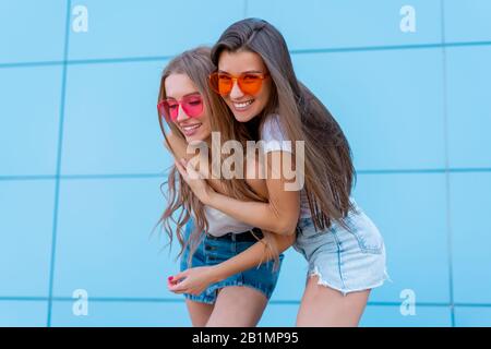 Due giovani hipster donna amici in neon retrò occhiali da sole in piedi e sorridente sulla parete blu Foto Stock