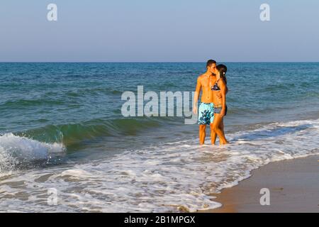 Abbracciando la giovane coppia baciata in pantaloncini beachwear godendo romantica passeggiata a piedi nudi sulla spiaggia di sabbia al tramonto. Stile di vita di viaggio concetto di vacanza. Foto Stock