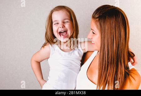 Sorridente giovane e informale femmina redhead che tiene ridendo piccola figlia in bianco tank top, mentre si diverte insieme in piedi su sfondo chiaro Foto Stock