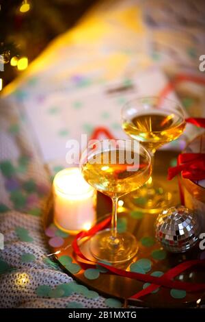 Elegante regalo e calici con champagne posto vicino a candela e piccola palla da discoteca sul vassoio su letto morbido durante la festa di Natale a casa Foto Stock