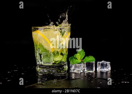 cocktail mojito con menta e lime al limone in un bicchiere di spruzzaggio con cubetti di ghiaccio, gocce e schizzi su sfondo nero Foto Stock