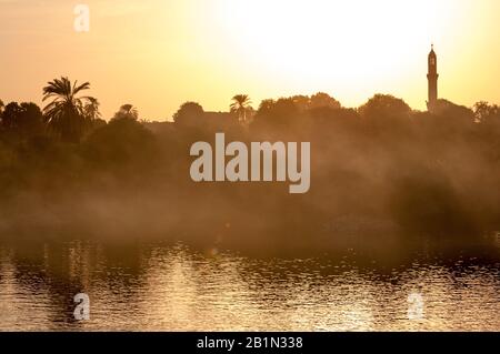 Golden sunlit vista delle nebbiose rive del fiume Nilo con sagome di palme e minareto moschea in Egitto Foto Stock