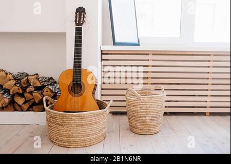 Chitarra acustica arancione di legno in un cesto a maglia sullo sfondo di un camino artificiale e tronchi. Una sala da musicista in stile boho. Foto Stock