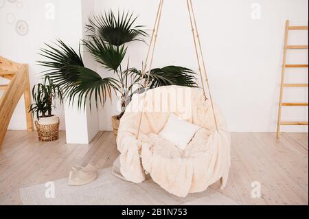 Appendete l'altalena alle corde nella stanza di casa con peluche plaid beige e cuscini. Un luogo dove rilassarsi e rilassarsi a casa in uno stile boho naturale Foto Stock