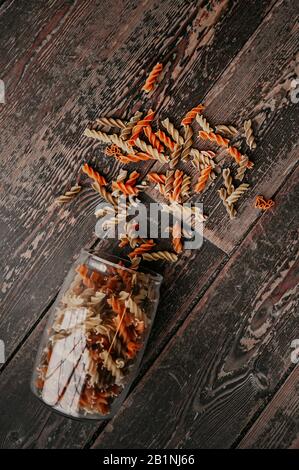 pasta cruda multicolore, arancione e gialla, sparsa da un vaso di vetro su un pavimento di legno scuro Foto Stock