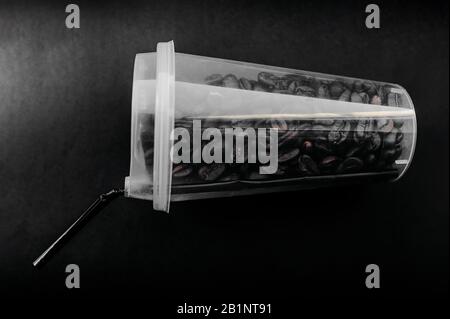 elegante vetro alto trasparente con una cannuccia nera piena di chicchi di caffè crudi su uno sfondo piatto denso nello stile di nero totale minimale, stylis Foto Stock