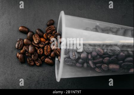 i chicchi di caffè arrostiti si riversano dal vetro trasparente in plastica su uno sfondo uniforme e denso nello stile di un caffè nero elegante e minimalista Foto Stock