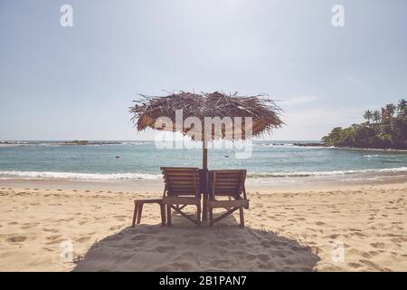 Lettini in legno sotto l'ombrello di palma su una spiaggia tropicale, tonificante colore applicato. Foto Stock