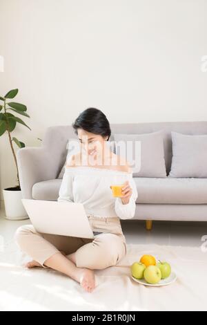 Bella donna asiatica utilizzando computer portatile mentre è seduto sul pavimento e di bere succo di frutta Foto Stock