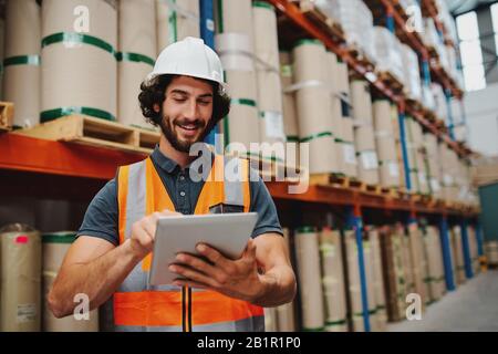 Responsabile del magazzino maschile che aggiunge i dati di inventario delle scorte in tablet digitali in magazzino indossando un elmetto bianco e un giubbotto di sicurezza in piedi Foto Stock