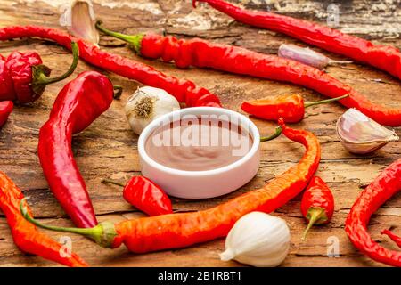 Salsa tradizionale Thai Sriracha con ingredienti. Peperoncino rosso e arancio leggermente secco, aglio. In ciotola di ceramica, sfondo di corteccia di legno, primo piano Foto Stock