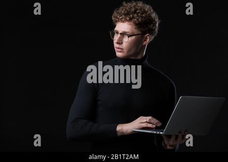 Bel sicuro giovane uomo d'affari riccioso indossando dolcevita in piedi davanti a sfondo isolato, lavorando su computer portatile Foto Stock