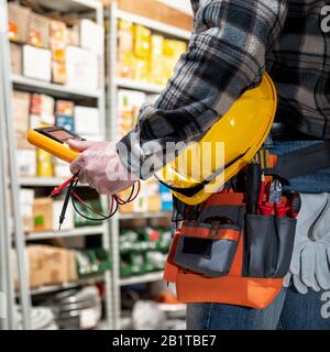 L'elettricista tiene a portata di mano il tester per multimetro, l'elmetto con occhiali di protezione. Industria edile, impianto elettrico. Foto Stock