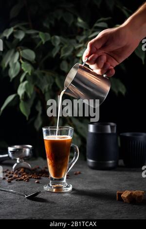 La mano maschile versa il latte da una caraffa in metallo in vetro trasparente con caffè nero su superficie scura e foglie di albero verde sullo sfondo Foto Stock