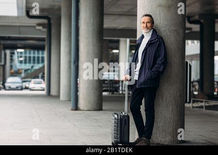 Elegante e maturo centro turistico all'aperto. Uomo con una valigia appoggiata sul muro che guarda lontano. Foto Stock