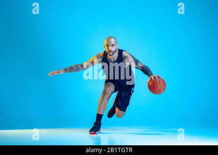 Giovane giocatore di basket della squadra che indossa l'allenamento sportivo, praticando in azione, movimento isolato su sfondo blu in luce al neon. Concetto di sport, movimento, energia e stile di vita dinamico e sano. Foto Stock