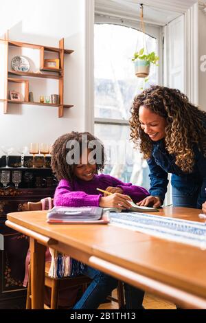 Madre sorridente che assiste la figlia nel fare i compiti a casa al tavolo nel soggiorno Foto Stock