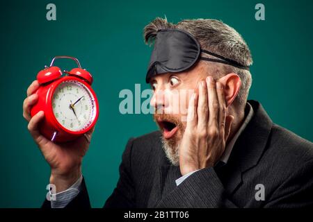 Businessman bearded in suit witn maschera di sonno sulla testa che tiene la sveglia rossa ed i yawns, vuole dormire. Concetto di gestione del tempo Foto Stock