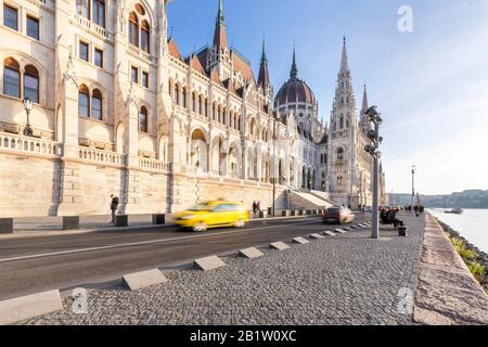 Budapest, Ungheria - 26 novembre 2019: Edificio del parlamento ungherese di fronte e un taxi che passa sulla riva del fiume in una giornata di sole nella stagione autunnale. Foto Stock