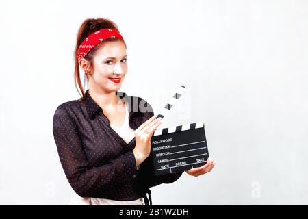 Giovane redhead felice pin up ragazza in bandana e camicia nera con labbra rosse sorridenti e tenendo il film clapper Foto Stock