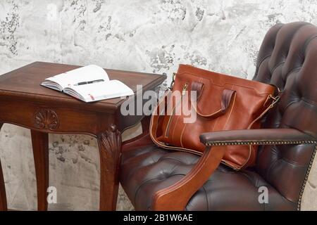 Borsa da lavoro in pelle e accessori nella sala da lavoro con parete in cemento grigio, tavolo in legno e sedia in pelle. Foto Stock