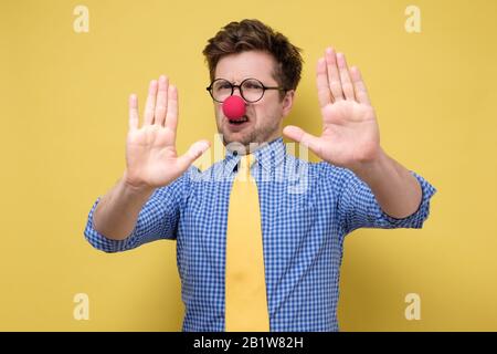 Divertente uomo caucasico con naso clown rosso su sfondo giallo isolato facendo un gesto di stop. Primo concetto di aprile Foto Stock